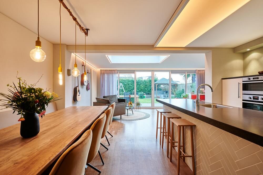 Plameco Spanndecke: Küche mit angrenzendem Wohnraum: Lichtmodul mit Human Centric Lighting über der Kücheninsel, Hängelampen über dem Esstisch und integrierter LED-Line am Deckenrand im Wohnbereich.